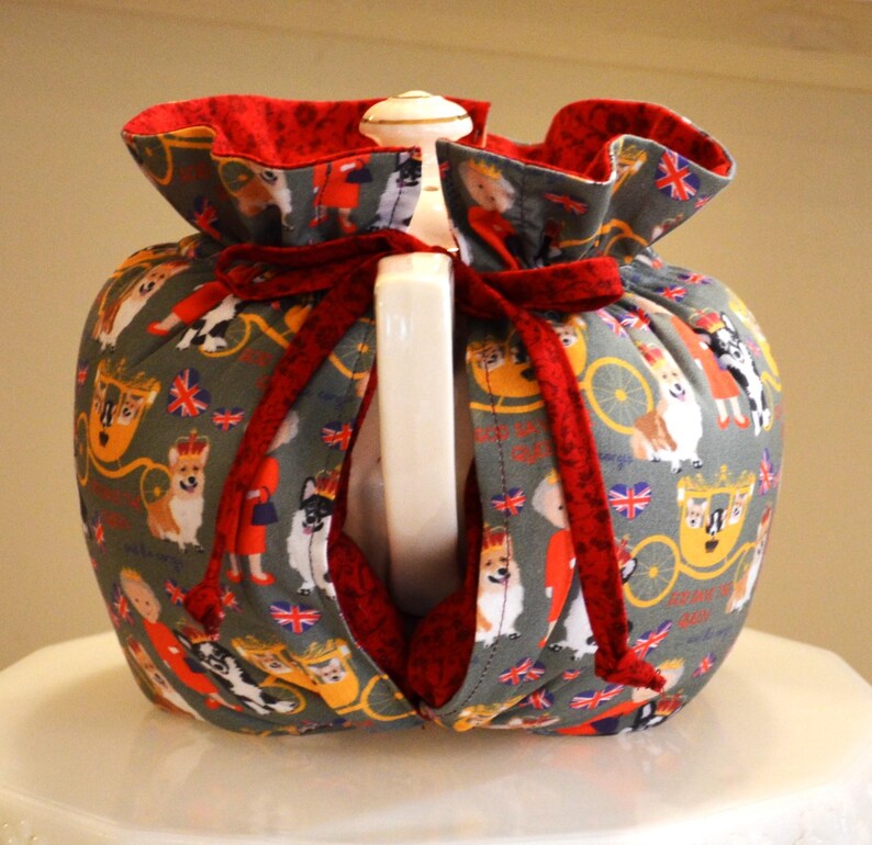 Puede incluir: Un calentador de t&eacute; gris con interior rojo y lazo rojo. El tejido presenta im&aacute;genes de perros corgi, coronas y carruajes. Envuelve una tetera blanca.