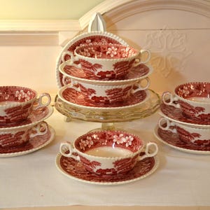 May include: A collection of vintage soup bowls and saucers. The bowls and saucers are white with a red floral and landscape pattern. The bowls have two handles. The set is arranged on a white surface.