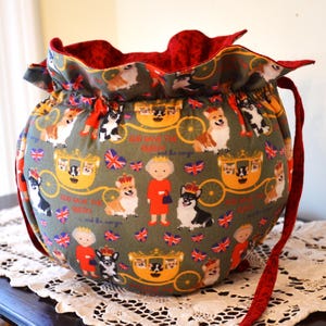 Puede incluir: Una bolsa redonda con cord&oacute;n y correa interior y roja. El exterior presenta un patr&oacute;n repetido de corgis, la Reina, carruajes y banderas de la Union Jack sobre un fondo verde. La bolsa est&aacute; sobre un mantel de encaje blanco.