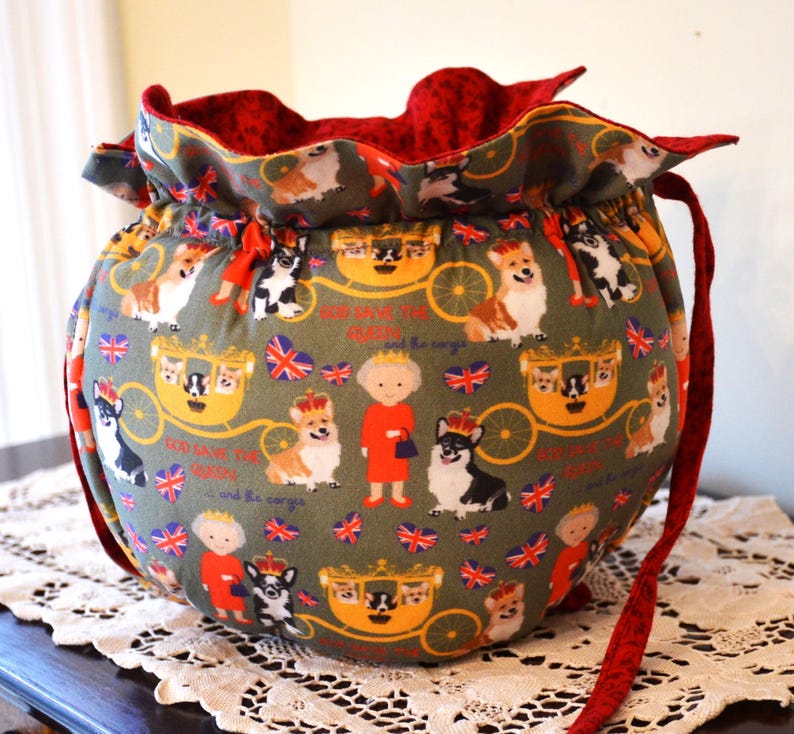 Puede incluir: Una bolsa redonda con cord&oacute;n y correa interior y roja. El exterior presenta un patr&oacute;n repetido de corgis, la Reina, carruajes y banderas de la Union Jack sobre un fondo verde. La bolsa est&aacute; sobre un mantel de encaje blanco.