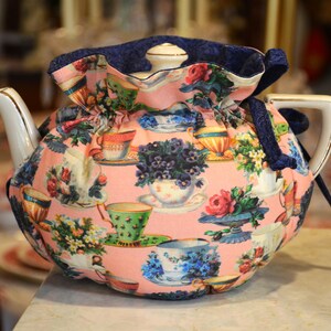 Puede incluir: Una tetera de cer&aacute;mica con un estampado floral y un fondo rosa. La tetera est&aacute; cubierta con una funda a juego con cierre de cord&oacute;n y ribete azul marino. La funda est&aacute; decorada con im&aacute;genes de tazas de t&eacute; y flores.