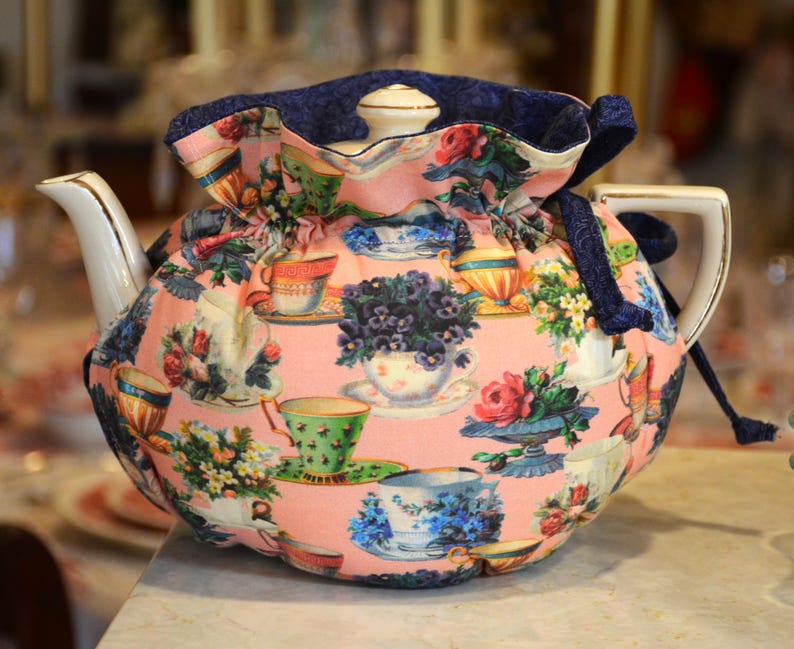 Puede incluir: Una tetera de cer&aacute;mica con un estampado floral y un fondo rosa. La tetera est&aacute; cubierta con una funda a juego con cierre de cord&oacute;n y ribete azul marino. La funda est&aacute; decorada con im&aacute;genes de tazas de t&eacute; y flores.