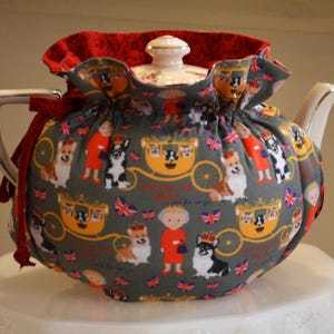 Puede incluir: Una tetera de cer&aacute;mica con una funda decorativa. La funda gris presenta un estampado de corgis, figuras reales y carrozas. Tiene un ribete rojo con volantes y un lazo rojo. La tetera tiene una tapa y un pico blancos.