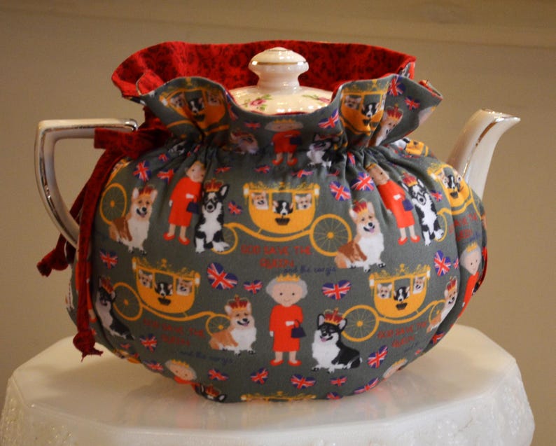 Puede incluir: Una tetera de cer&aacute;mica con una funda decorativa. La funda gris presenta un estampado de corgis, figuras reales y carrozas. Tiene un ribete rojo con volantes y un lazo rojo. La tetera tiene una tapa y un pico blancos.