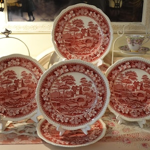 May include: A set of five ceramic plates featuring a red and white transferware pattern. Each plate displays a detailed landscape scene with a stone bridge, trees, and buildings, surrounded by a decorative floral border with scalloped edges.