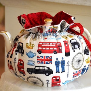 K&ouml;nnte beinhalten: Eine wei&szlig;e Teekanne mit einem roten und goldenen Teew&auml;rmer mit London-Themen-Illustrationen. Das Design umfasst rote Doppeldeckerbusse, schwarze Taxis und die Union Jack. Die Teekanne hat einen goldverzierten Ausguss und Griff.