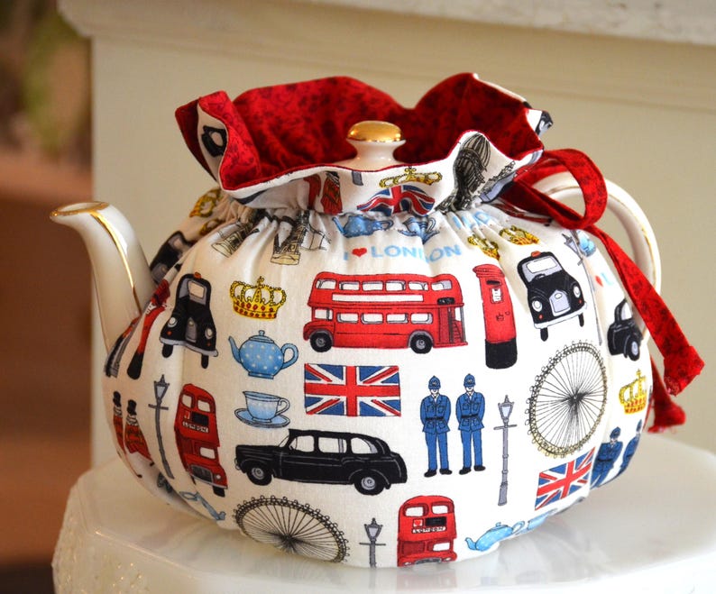 K&ouml;nnte beinhalten: Eine wei&szlig;e Teekanne mit einem roten und goldenen Teew&auml;rmer mit London-Themen-Illustrationen. Das Design umfasst rote Doppeldeckerbusse, schwarze Taxis und die Union Jack. Die Teekanne hat einen goldverzierten Ausguss und Griff.