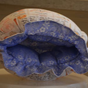 Może przedstawiać: Biało-niebieski materiałowy czepek z kwiatowym wzorem i zapięciem na sznurek. Wnętrze to jasnoniebieska tkanina z białym kwiatowym wzorem. Na zewnątrz znajdują się ilustracje jedzenia i napis.