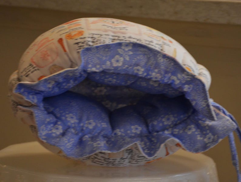 Może przedstawiać: Biało-niebieski materiałowy czepek z kwiatowym wzorem i zapięciem na sznurek. Wnętrze to jasnoniebieska tkanina z białym kwiatowym wzorem. Na zewnątrz znajdują się ilustracje jedzenia i napis.