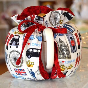 K&ouml;nnte beinhalten: Eine wei&szlig;e Teekanne mit roter Verzierung und Schleife. Die Teekanne ist mit London-Themen-Illustrationen verziert, darunter schwarze Taxis, das London Eye und der Union Jack. Die Teekanne hat einen wei&szlig;en Griff.