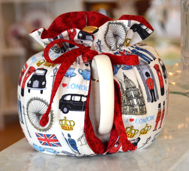 K&ouml;nnte beinhalten: Eine wei&szlig;e Teekanne mit roter Verzierung und Schleife. Die Teekanne ist mit London-Themen-Illustrationen verziert, darunter schwarze Taxis, das London Eye und der Union Jack. Die Teekanne hat einen wei&szlig;en Griff.