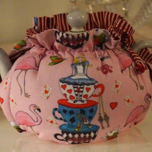 May include: A pink teapot cozy with an Alice in Wonderland theme. The cozy features illustrations of flamingos, playing cards, and Alice standing on teacups. The top is trimmed with red and white striped fabric.
