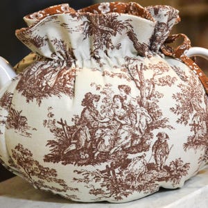 May include: A white ceramic teapot with a brown and white toile fabric cozy. The fabric features a pastoral scene with figures and trees. The cozy has a ruffled top and a drawstring closure. The handle and spout are white.