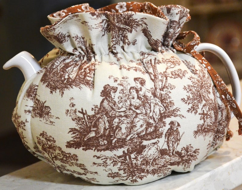 May include: A white ceramic teapot with a brown and white toile fabric cozy. The fabric features a pastoral scene with figures and trees. The cozy has a ruffled top and a drawstring closure. The handle and spout are white.
