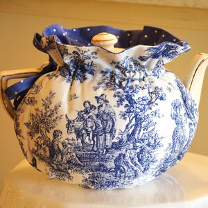 Op de afbeelding: Een witte keramische theepot met een blauw-wit toile de Jouy-patroon, met een pastorale sc&egrave;ne. De theepot is bedekt met een bijpassende stoffen theemuts met een blauwe stippelrand en een blauw lint. Het deksel heeft een kleine, ronde, cr&egrave;mekleurige knop.
