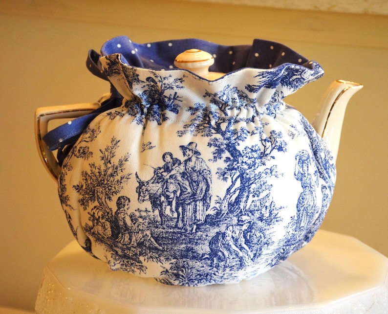 Op de afbeelding: Een witte keramische theepot met een blauw-wit toile de Jouy-patroon, met een pastorale sc&egrave;ne. De theepot is bedekt met een bijpassende stoffen theemuts met een blauwe stippelrand en een blauw lint. Het deksel heeft een kleine, ronde, cr&egrave;mekleurige knop.