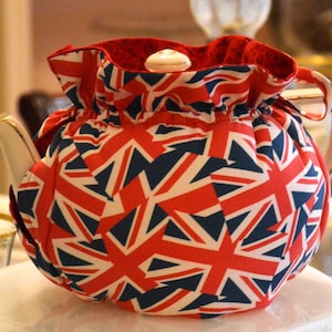 May include: A white porcelain teapot with a red, white, and blue Union Jack patterned cozy. The cozy has a ruffled edge and covers the teapot. The teapot has a gold handle and spout. A small white lid sits on top.