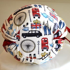 K&ouml;nnte beinhalten: Ein wei&szlig;er Teew&auml;rmer mit einem London-Themenmuster. Das Design umfasst rote Doppeldeckerbusse, schwarze Taxis, den Union Jack und das London Eye. Der W&auml;rmer hat rote Verzierungen und einen Griff.