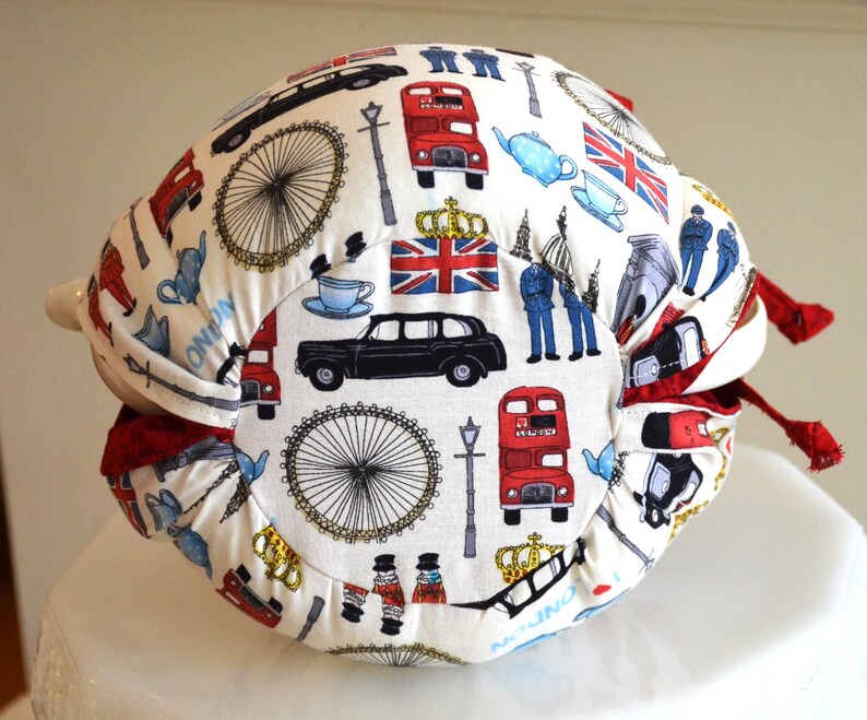 K&ouml;nnte beinhalten: Ein wei&szlig;er Teew&auml;rmer mit einem London-Themenmuster. Das Design umfasst rote Doppeldeckerbusse, schwarze Taxis, den Union Jack und das London Eye. Der W&auml;rmer hat rote Verzierungen und einen Griff.