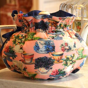 Puede incluir: Una tetera de porcelana blanca con asa y boquilla doradas, cubierta por un cubre tetera rosa. El cubre tetera est&aacute; estampado con tazas de t&eacute;, flores y arreglos florales en varios colores. Un cord&oacute;n azul marino asegura el cubre tetera.