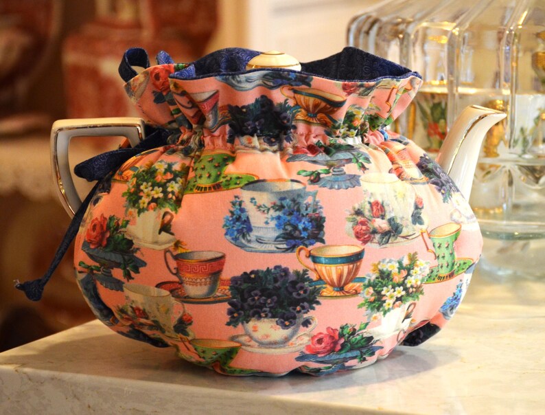 Puede incluir: Una tetera de porcelana blanca con asa y boquilla doradas, cubierta por un cubre tetera rosa. El cubre tetera est&aacute; estampado con tazas de t&eacute;, flores y arreglos florales en varios colores. Un cord&oacute;n azul marino asegura el cubre tetera.