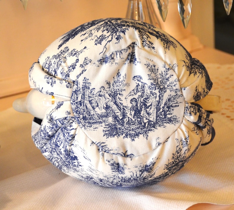 Op de afbeelding: Een wit en blauw toile de Jouy wijnfleshoes. De stof heeft een pastorale sc&egrave;ne met figuren en bomen. De hoes is om een fles gewikkeld, waarbij een deel van de hals zichtbaar is.