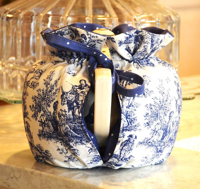 Op de afbeelding: Een wit en blauw toilettasje met een blauw lint. De tas heeft een klassiek toile-patroon met figuren en bomen. Een wit rechthoekig object is zichtbaar in de tas.