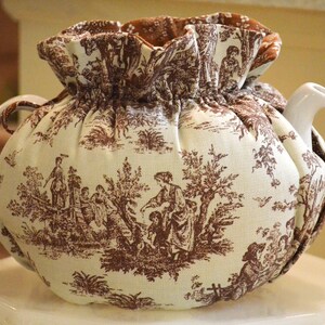 May include: A white ceramic teapot with a brown and white toile tea cozy. The cozy features a pastoral scene with figures and trees. The cozy has a ruffled top and drawstring closure. The teapot has a curved handle and spout.