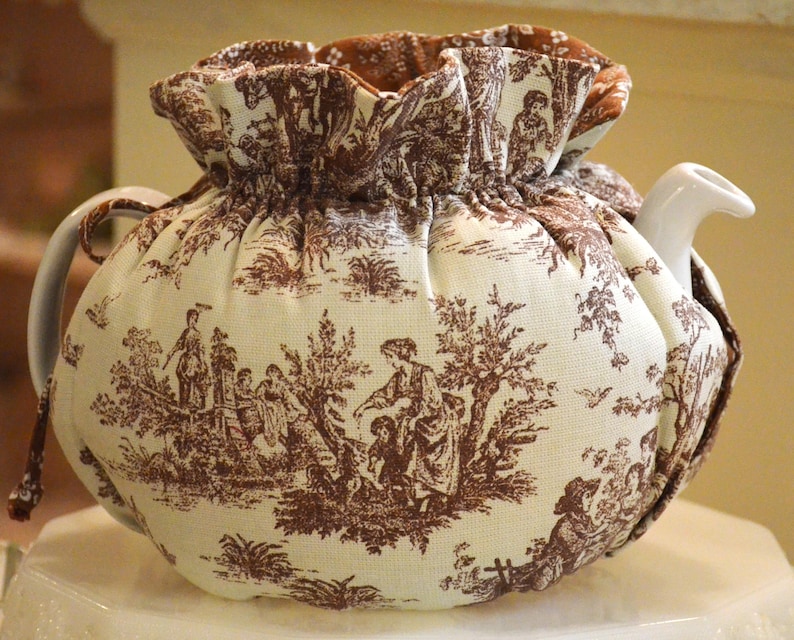 May include: A white ceramic teapot with a brown and white toile tea cozy. The cozy features a pastoral scene with figures and trees. The cozy has a ruffled top and drawstring closure. The teapot has a curved handle and spout.