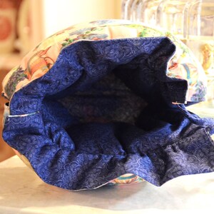 Puede incluir: Una bolsa con cord&oacute;n con un estampado floral y de p&aacute;jaros en el exterior y un forro azul oscuro estampado. La abertura de la bolsa est&aacute; fruncida, revelando la tela interior azul. La bolsa est&aacute; sobre una superficie clara.