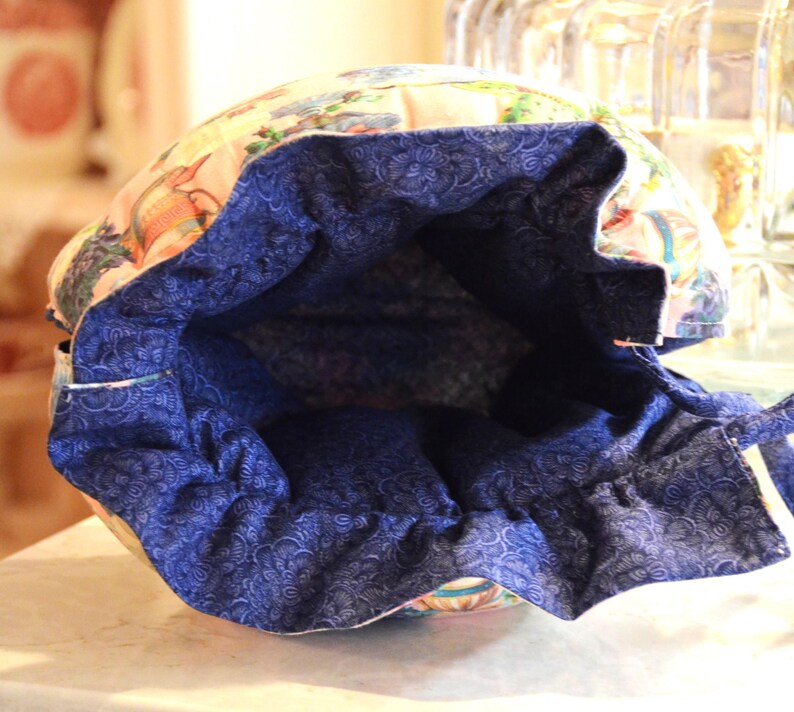 Puede incluir: Una bolsa con cord&oacute;n con un estampado floral y de p&aacute;jaros en el exterior y un forro azul oscuro estampado. La abertura de la bolsa est&aacute; fruncida, revelando la tela interior azul. La bolsa est&aacute; sobre una superficie clara.