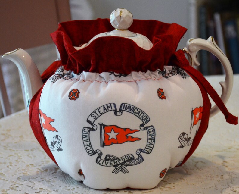 Puede incluir: Tetera blanca con una cubierta de tela roja. La cubierta tiene un logotipo azul marino que dice "Oceanic Steam Navigation Company Liverpool" con una estrella roja y una bandera roja.