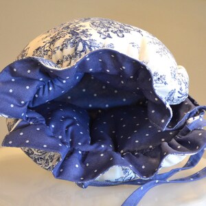 Op de afbeelding: Een blauw-witte bonnet met een bloemenpatroon en een gerimpelde rand. De binnenkant van de rand is effen blauw met witte stippen. De bonnet heeft blauwe strikjes.