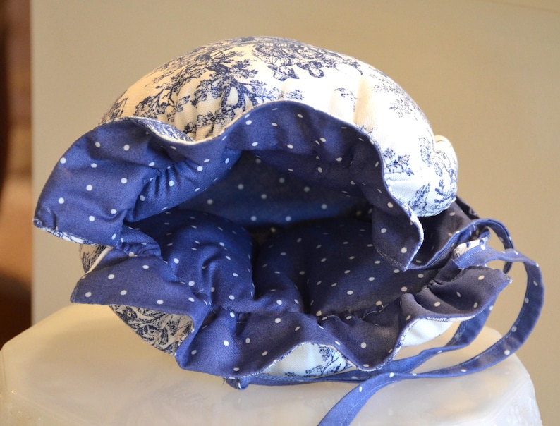 Op de afbeelding: Een blauw-witte bonnet met een bloemenpatroon en een gerimpelde rand. De binnenkant van de rand is effen blauw met witte stippen. De bonnet heeft blauwe strikjes.