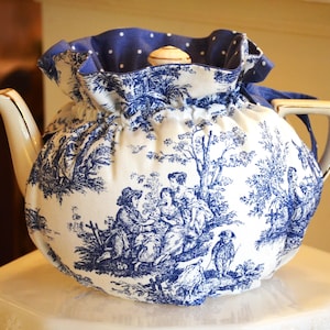 Op de afbeelding: Een witte porseleinen theepot met een blauw-wit toile de Jouy-patroon, bedekt met een bijpassende theemuts. De theemuts heeft een blauwe stippenvoering en een trekkoordsluiting. De theepot heeft een gouden handvat en tuit.
