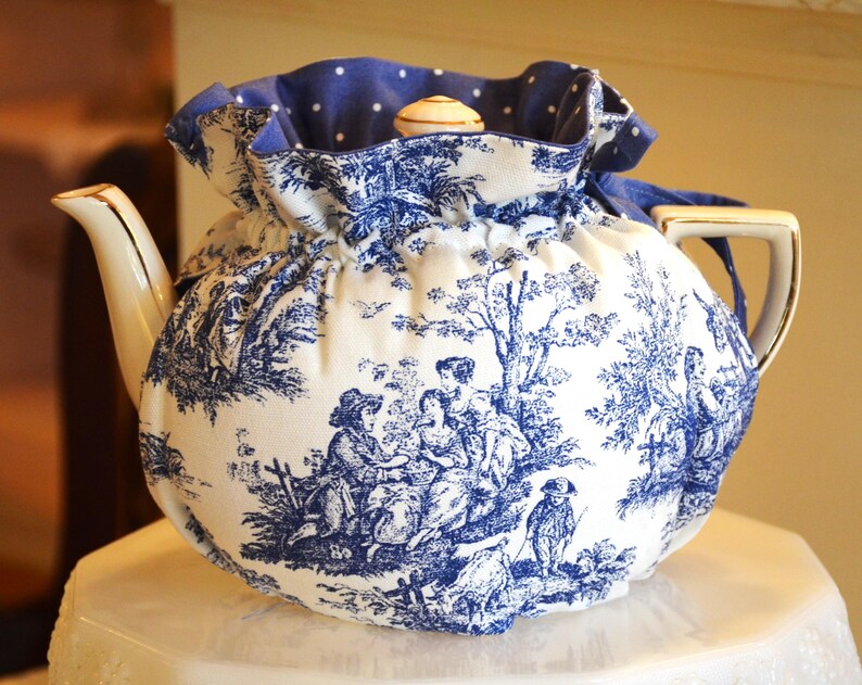 Op de afbeelding: Een witte porseleinen theepot met een blauw-wit toile de Jouy-patroon, bedekt met een bijpassende theemuts. De theemuts heeft een blauwe stippenvoering en een trekkoordsluiting. De theepot heeft een gouden handvat en tuit.