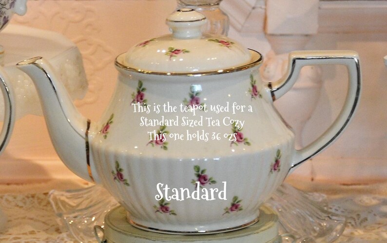 Op de afbeelding: Een witte porseleinen theepot met gouden rand en deksel, versierd met roze rozenpatronen. De theepot heeft een gebogen tuit en een handvat. De tekst op de theepot luidt "Standard" en "This one holds 36 oz".