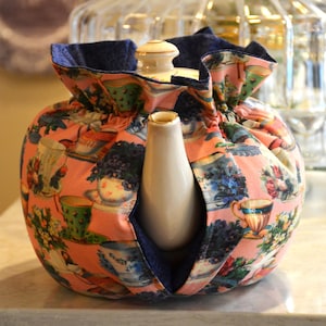 Puede incluir: Un cubreteiera rosa con un estampado floral y de tazas de t&eacute;. El cubreteiera tiene un forro azul marino y est&aacute; envuelto alrededor de una tetera blanca. Un accesorio de cocina decorativo para mantener la tetera caliente.