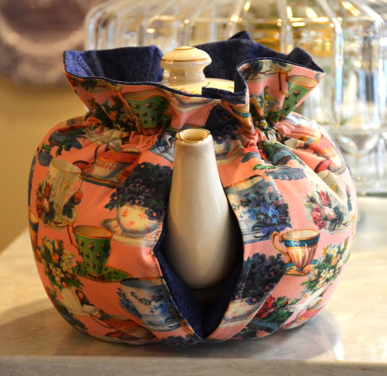 Puede incluir: Un cubreteiera rosa con un estampado floral y de tazas de t&eacute;. El cubreteiera tiene un forro azul marino y est&aacute; envuelto alrededor de una tetera blanca. Un accesorio de cocina decorativo para mantener la tetera caliente.