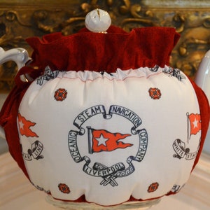 Puede incluir: Tetera blanca con un calienta-te rojo y blanco. El calienta-te tiene un volante rojo y presenta una estrella blanca sobre una bandera roja con el texto "Oceanic Steam Navigation Company Liverpool" en un c&iacute;rculo.