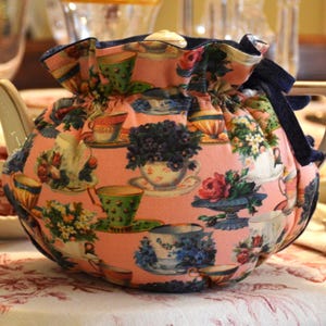 Puede incluir: Una tetera de cer&aacute;mica con una funda estampada con flores. La funda es rosa con im&aacute;genes de tazas de t&eacute;, flores y teteras. La tetera tiene un pico y un asa blancos con ribete dorado. Una cinta azul oscuro est&aacute; atada en la parte superior.