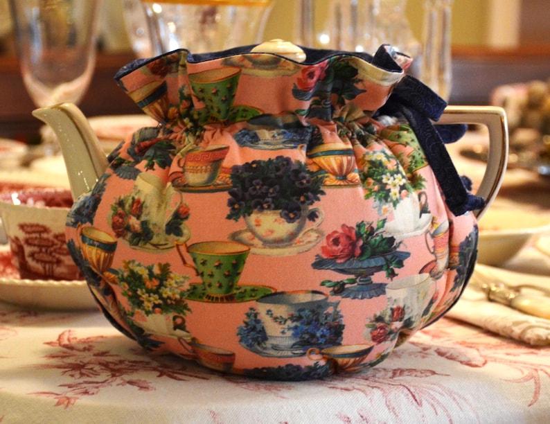 Puede incluir: Una tetera de cer&aacute;mica con una funda estampada con flores. La funda es rosa con im&aacute;genes de tazas de t&eacute;, flores y teteras. La tetera tiene un pico y un asa blancos con ribete dorado. Una cinta azul oscuro est&aacute; atada en la parte superior.