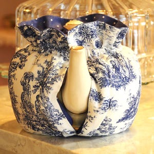 Op de afbeelding: Een witte keramische theepot met een blauw-witte toile de Jouy stoffen hoes. De stof toont een pastorale sc&egrave;ne met bomen en figuren. De hoes heeft een blauwe stippelrand en bedekt de theepot.