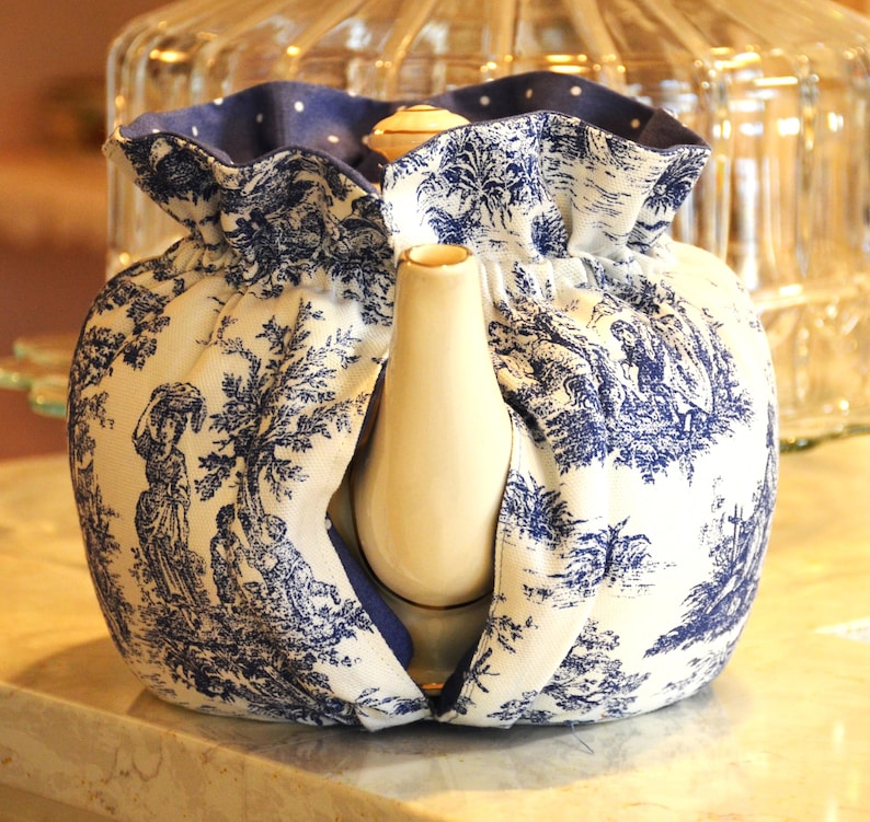 Op de afbeelding: Een witte keramische theepot met een blauw-witte toile de Jouy stoffen hoes. De stof toont een pastorale sc&egrave;ne met bomen en figuren. De hoes heeft een blauwe stippelrand en bedekt de theepot.