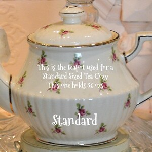 Puede incluir: Una tetera de porcelana blanca con ribete dorado y tapa, adornada con peque&ntilde;as rosas rosas y hojas verdes. El texto de la tetera dice: "This is the teapot used for a Standard Sized Tea Cozy. This one holds 36 oz."