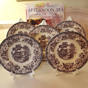 May include: Five white and brown decorative plates with a floral and landscape design are displayed on white plate stands. The plates have a scalloped edge. A cookbook titled "The Perfect Afternoon Tea Recipe Book" is in the background.