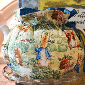 May include: A teapot cozy featuring a whimsical Peter Rabbit design. The fabric depicts scenes with rabbits, squirrels, and vegetables in a garden setting. The cozy is tied with a blue ribbon and sits on a clear glass plate.