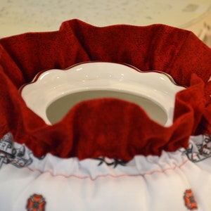 Puede incluir: Una tetera blanca con una cubierta de tela roja. La cubierta tiene un borde blanco y una cinta roja. La tetera tiene un borde dorado y un asa dorada.