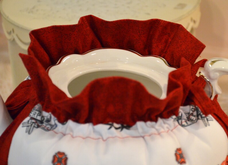 Puede incluir: Una tetera blanca con una cubierta de tela roja. La cubierta tiene un borde blanco y una cinta roja. La tetera tiene un borde dorado y un asa dorada.