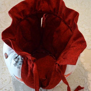 Puede incluir: Una bolsa de tela roja con cord&oacute;n de ajuste, forro blanco y base de tela blanca. La bolsa tiene un borde superior con volantes y un cierre de cord&oacute;n. La base de la bolsa tiene un patr&oacute;n de rayas blancas y rojas.